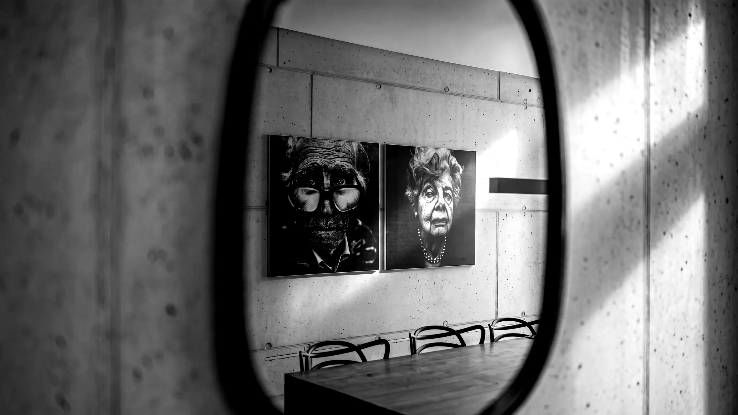 Mur de concept dans les bureaux de MOREMEDIA® : deux portraits noir et blanc grand format sur un mur en béton brut, encadrés par un miroir ovale au premier plan – table de réunion en chêne avec chaises.
