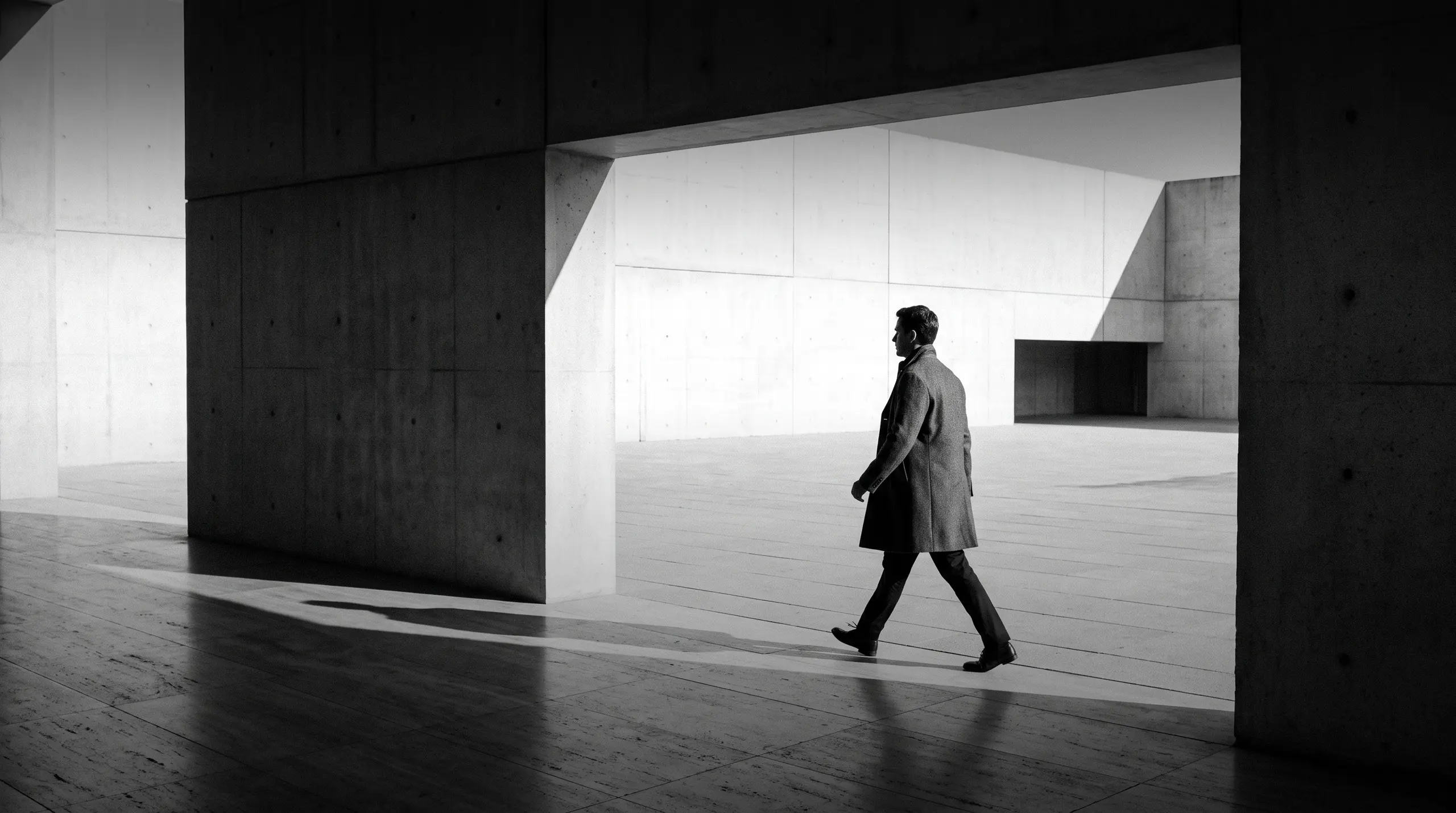 HUMAN STRUCTURES : Un homme marche de profil à travers une ouverture en béton, à contre-jour, passant d'un couloir sombre à une cour lumineuse.