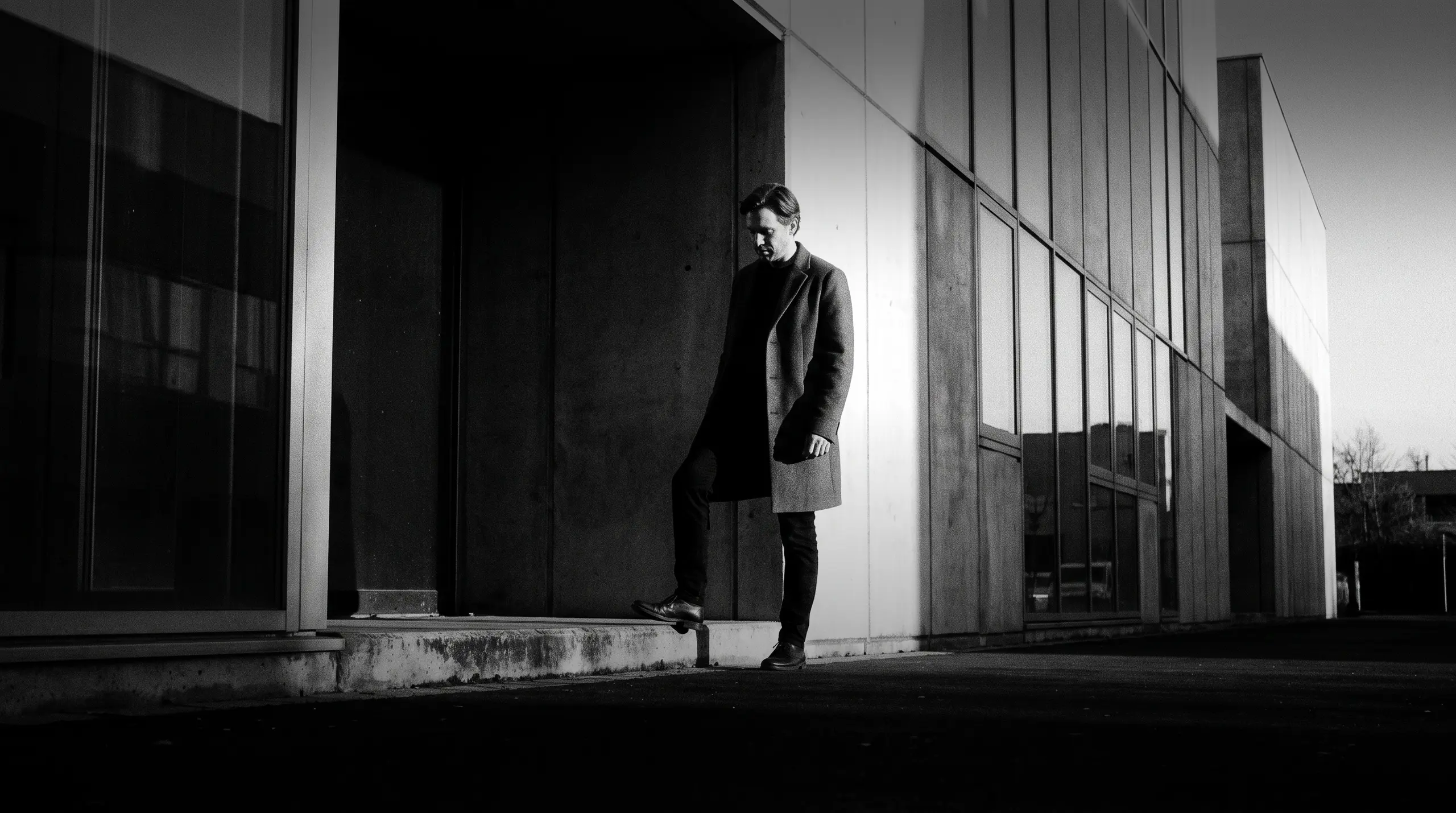 HUMAN STRUCTURES: Un homme en manteau pose un pied sur un rebord de béton à l'entrée sombre d'un bâtiment, le regard baissé.