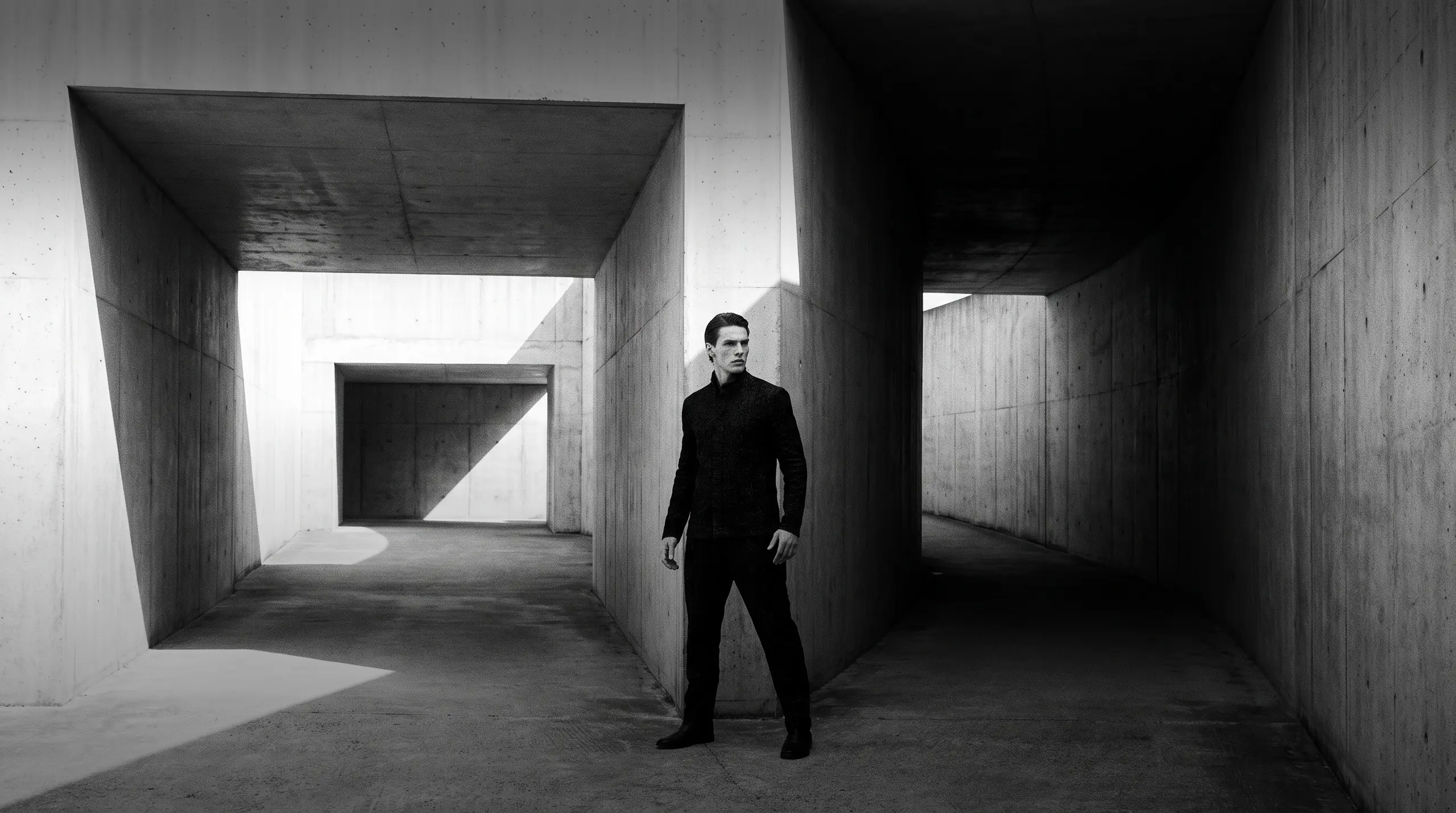 HUMAN STRUCTURES: Un homme vêtu de noir se tient seul au croisement d'étroits couloirs de béton, le regard de côté, effleuré par une fine bande de lumière.