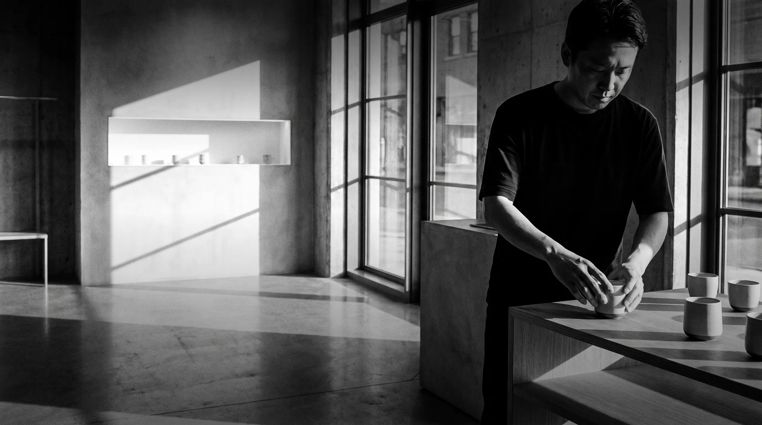 HUMAN STRUCTURES : Un homme façonne avec attention un bol en céramique sur un comptoir en bois, entouré de tasses alignées dans un intérieur en béton lumineux.