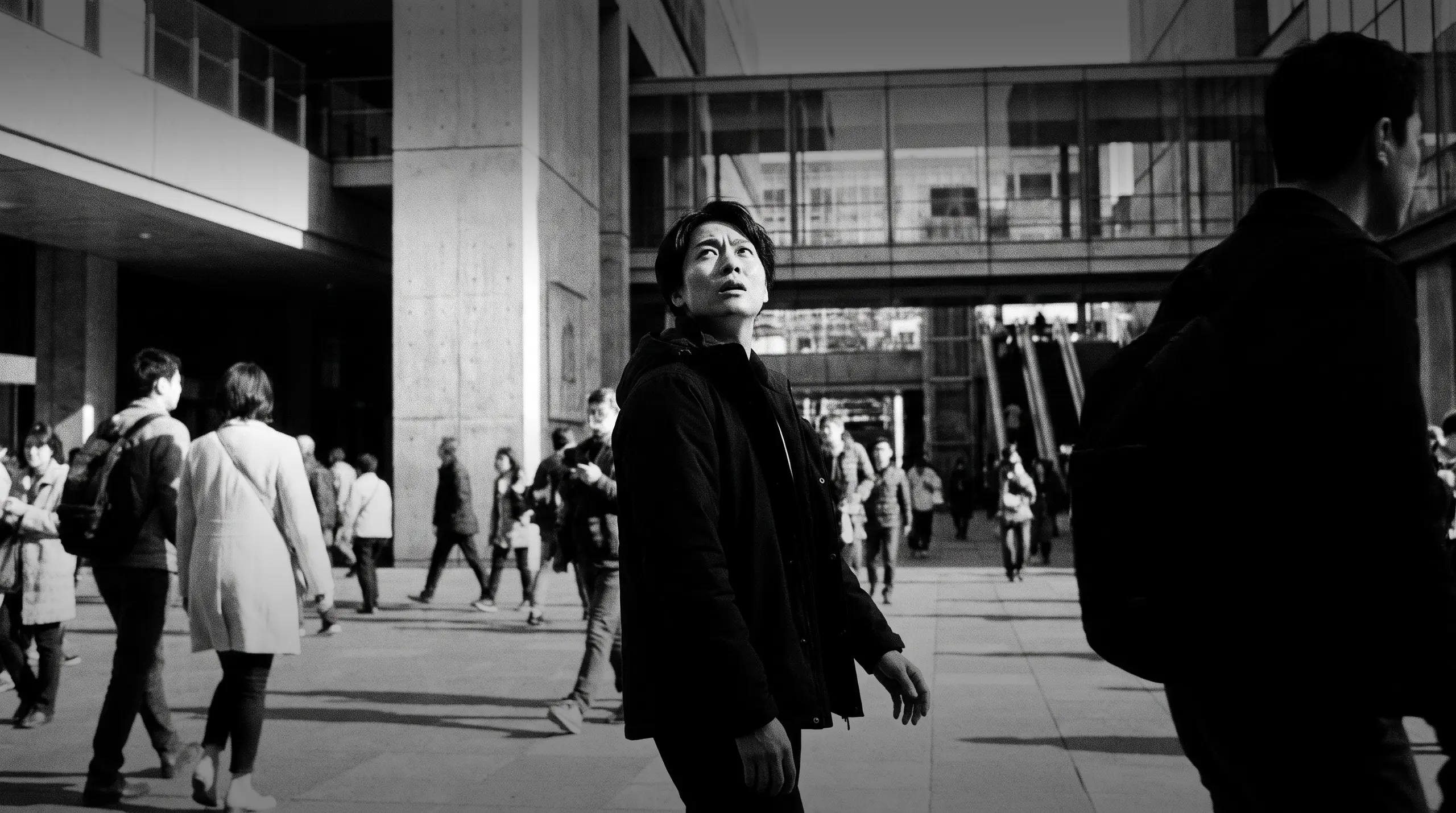 HUMAN STRUCTURES: Un jeune homme lève les yeux d'un air interrogateur tandis qu'une foule se déplace autour de lui sur une place bordée de façades vitrées.