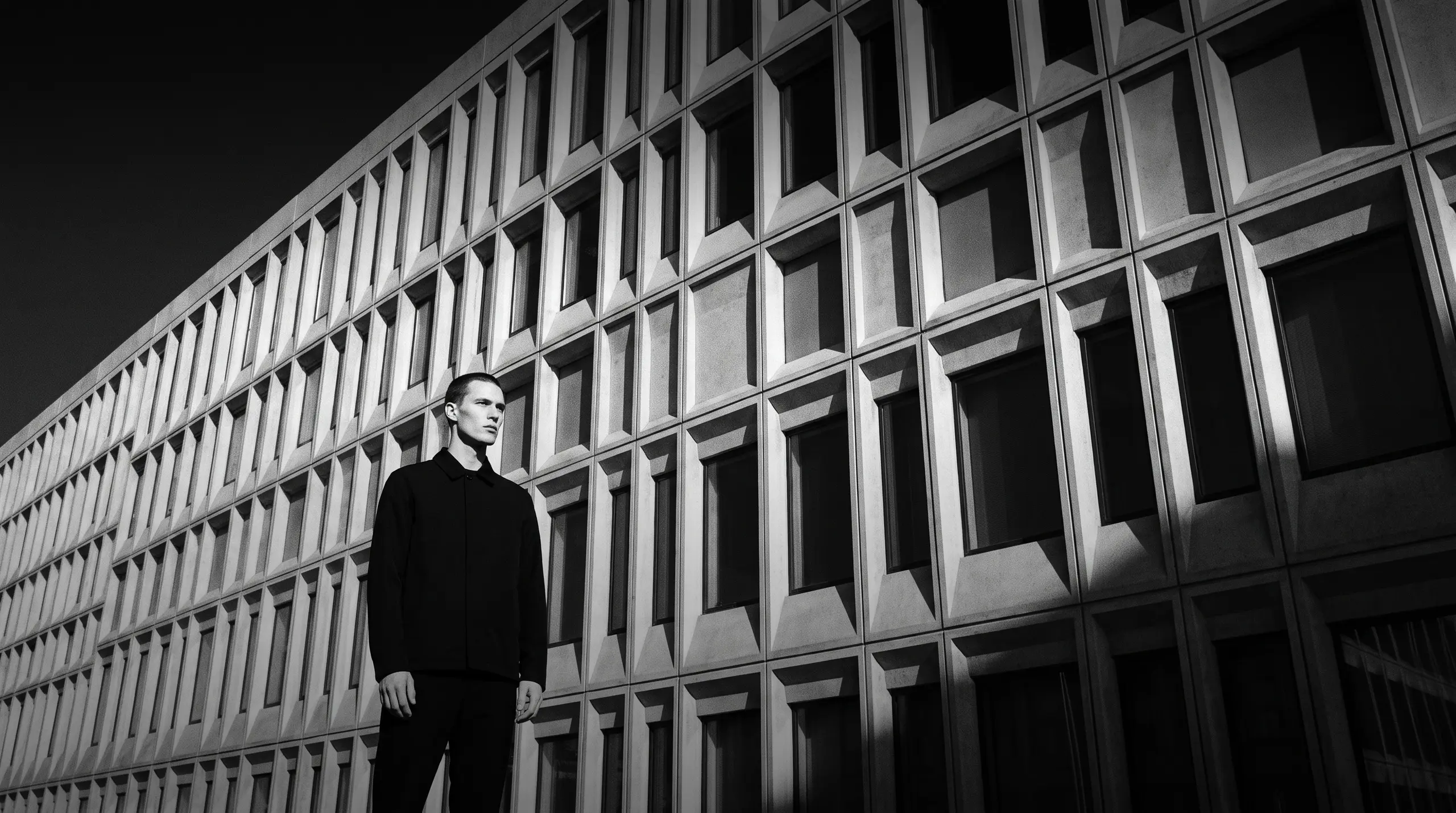 HUMAN STRUCTURES: Un homme paraît minuscule devant une immense façade de béton en gradins de lumière, aux niches de fenêtres disposées en symétrie stricte.