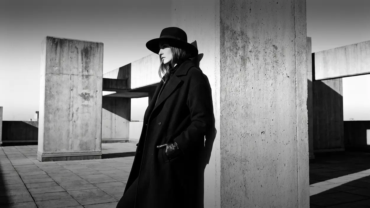 HUMAN STRUCTURES: A woman in a hat and long coat leans against a massive concrete pillar on a rooftop terrace, profile lit by hard sunlight.