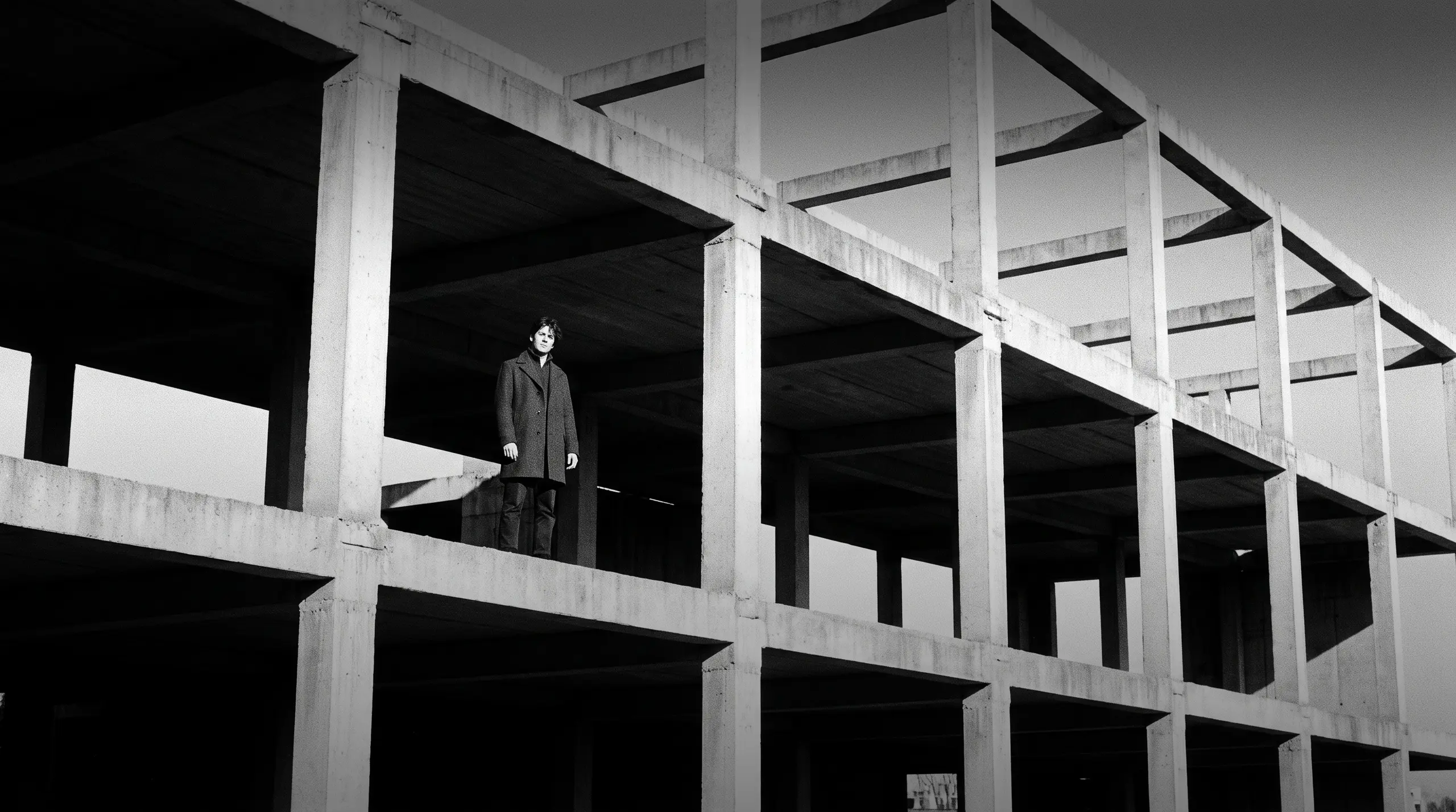 HUMAN STRUCTURES: A man in a dark coat stands on an open concrete level of a grid-like unfinished building, gazing into the distance.