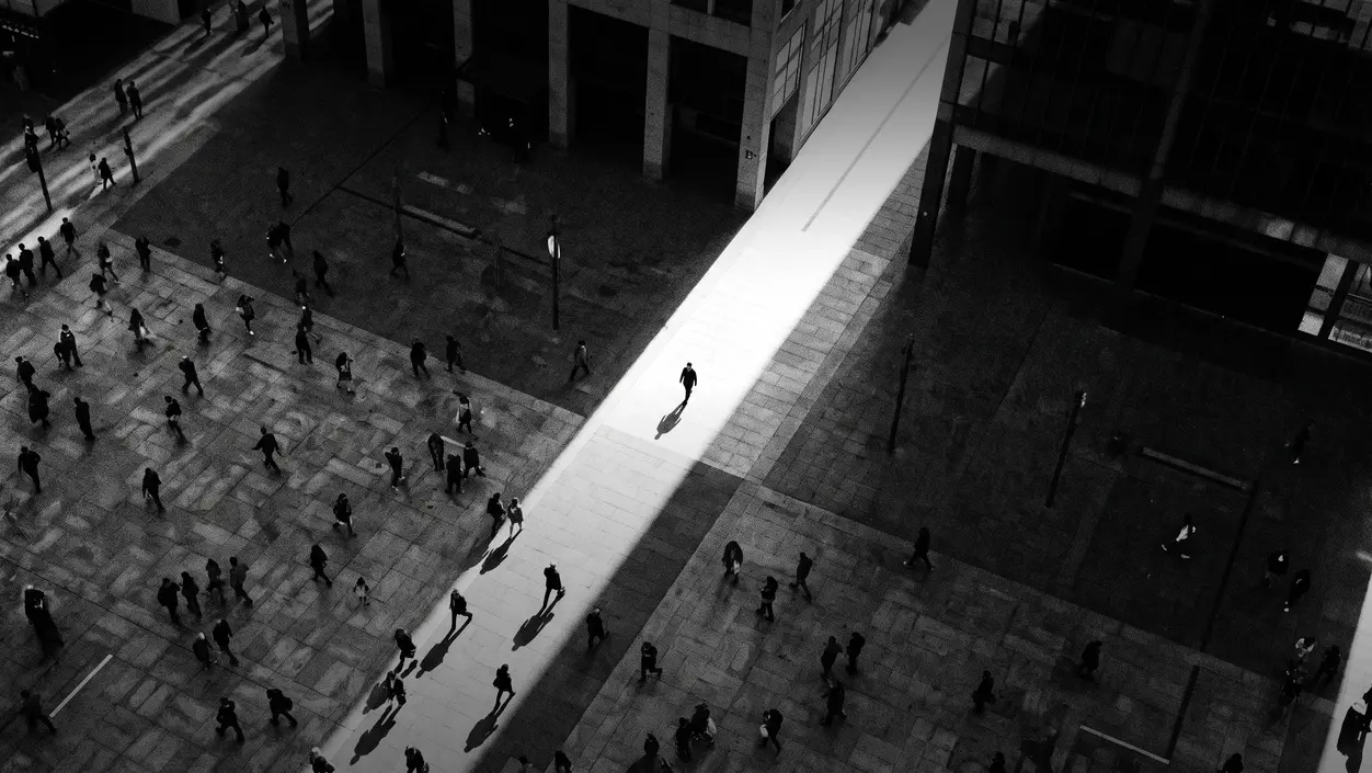 HUMAN STRUCTURES: Seen from above, one lone figure crosses a broad bright shaft of light amid darker passersby on a concrete square.
