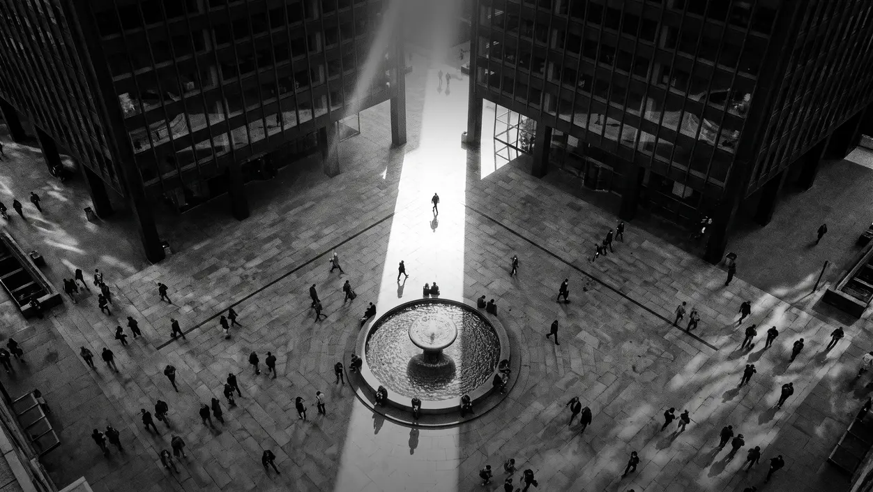 HUMAN STRUCTURES: Seen from above, a crowd crosses a plaza between two office towers, a bright shaft of light striking a round fountain at its center.
