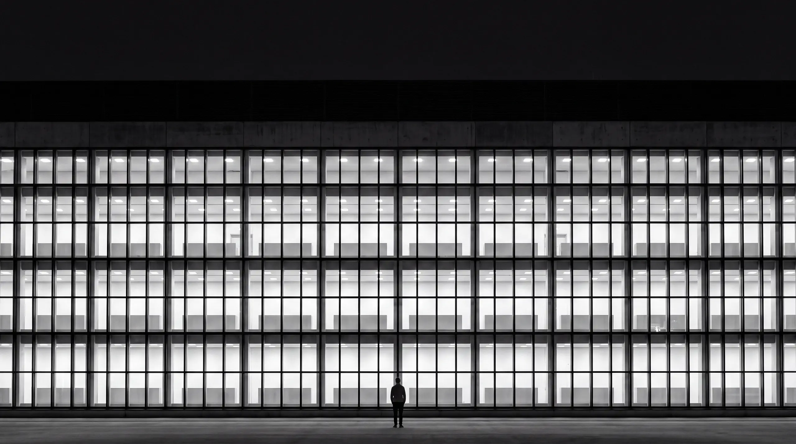 AI-generated image from the HUMAN STRUCTURES series: At dusk, a person stands in front of a long, strictly gridded glass façade with every window evenly lit from within.