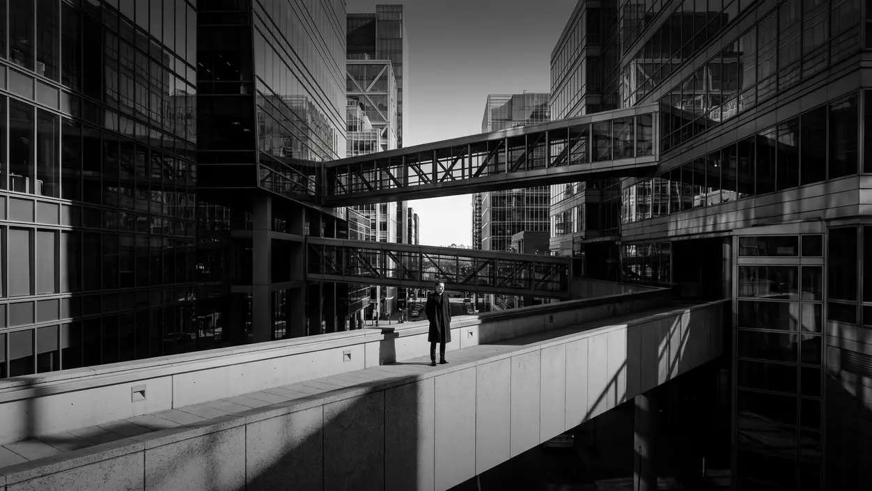 HUMAN STRUCTURES: A man in a dark coat stands on an elevated concrete walkway between glass office towers, flanked by skybridges and glass facades.